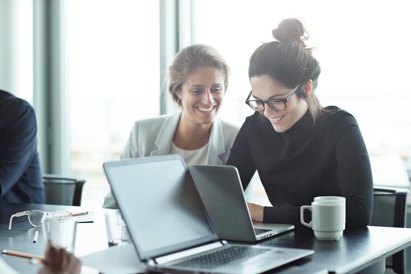 Två kvinnor sitter framför en laptop vid ett konferensbord och ler